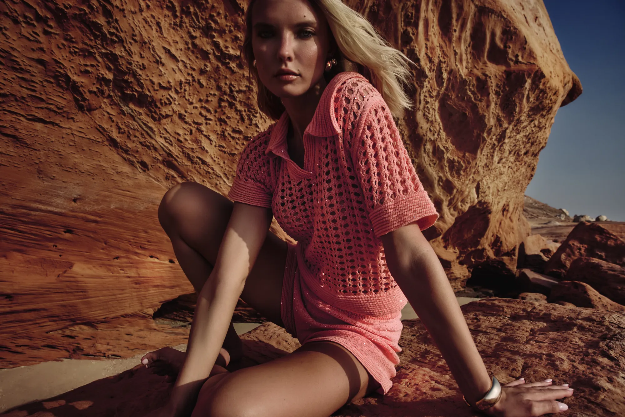 Model in a coral open-knit polo set crouched on sun-warmed red rock, gazing into the lens