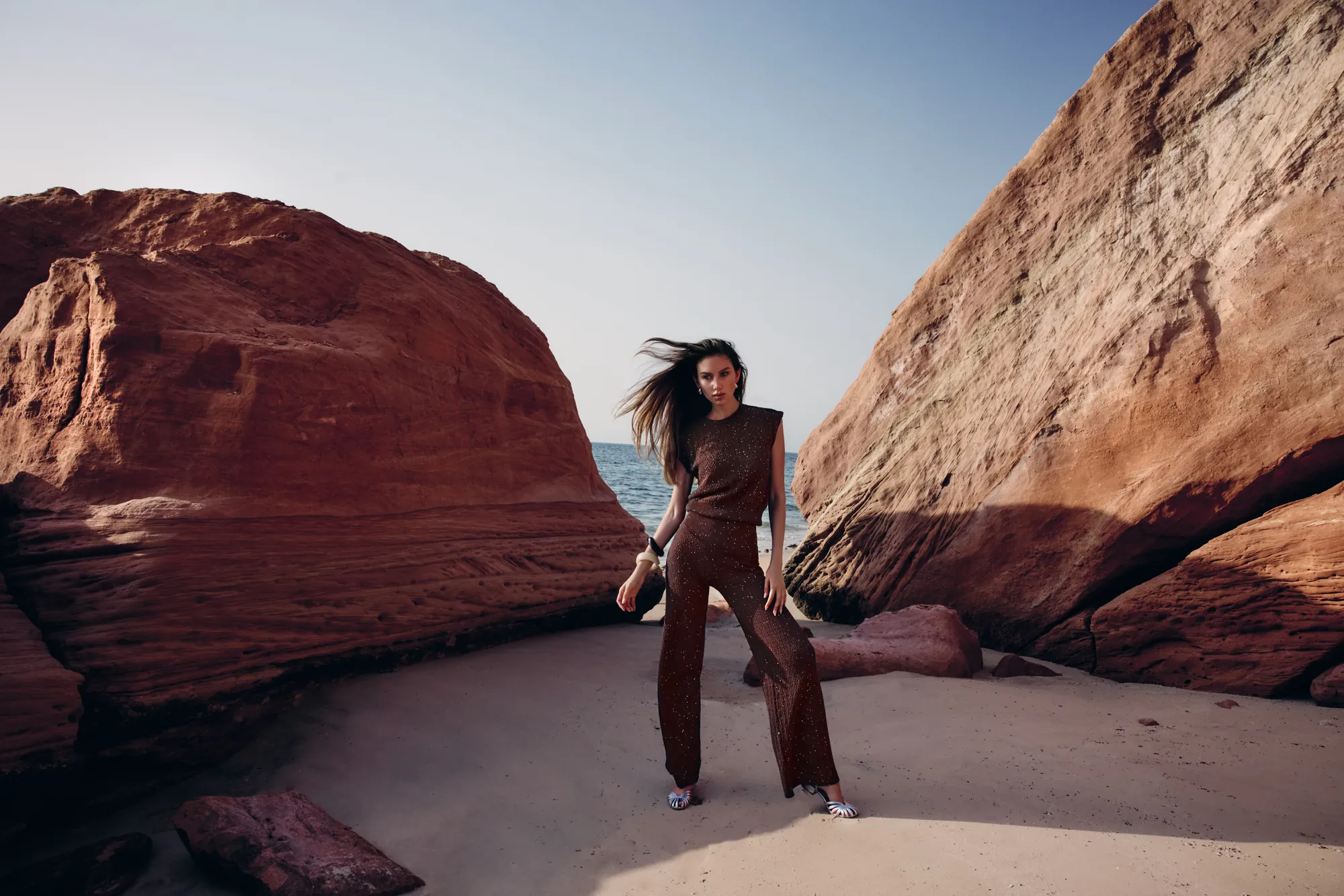 Model striding across pale sand in a chocolate knit co-ord, red monoliths on either side