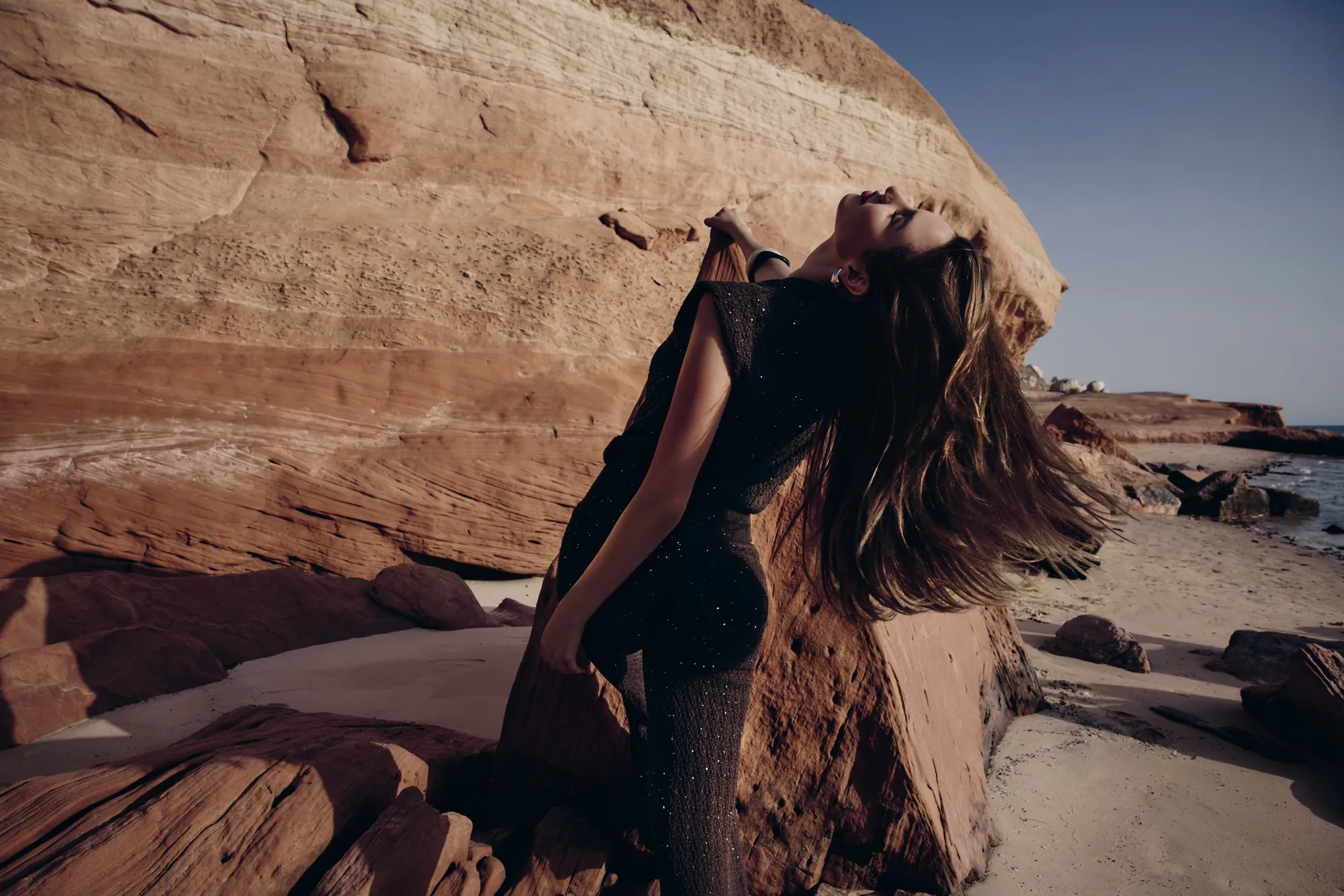 Model in a black sparkle-knit jumpsuit arching back against striated red cliffs at dusk