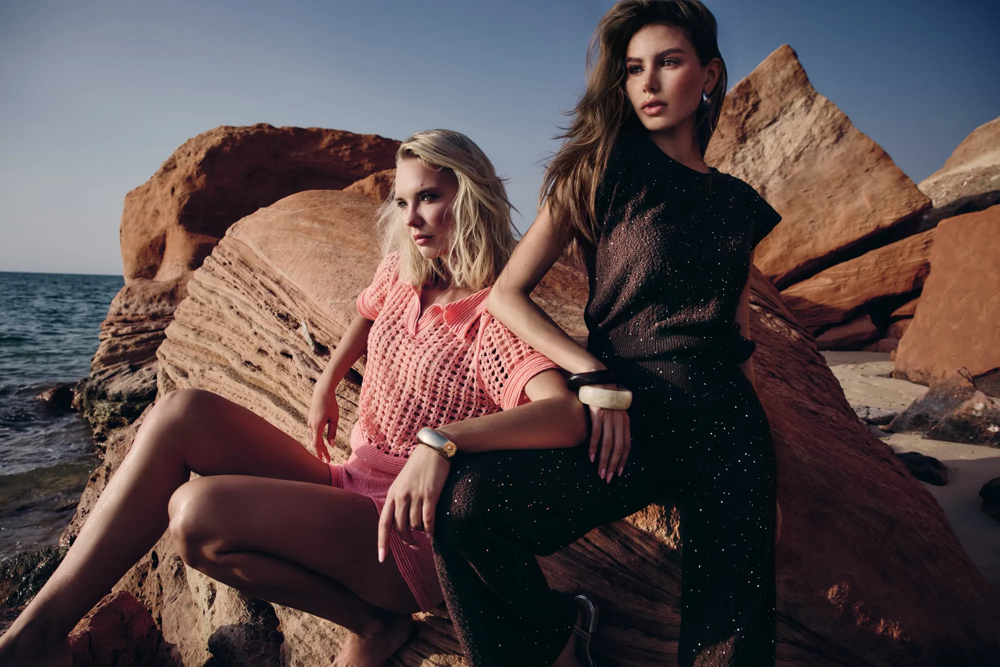 Two models in coral crochet and black sparkle knits perched on red rocks at the sea's edge