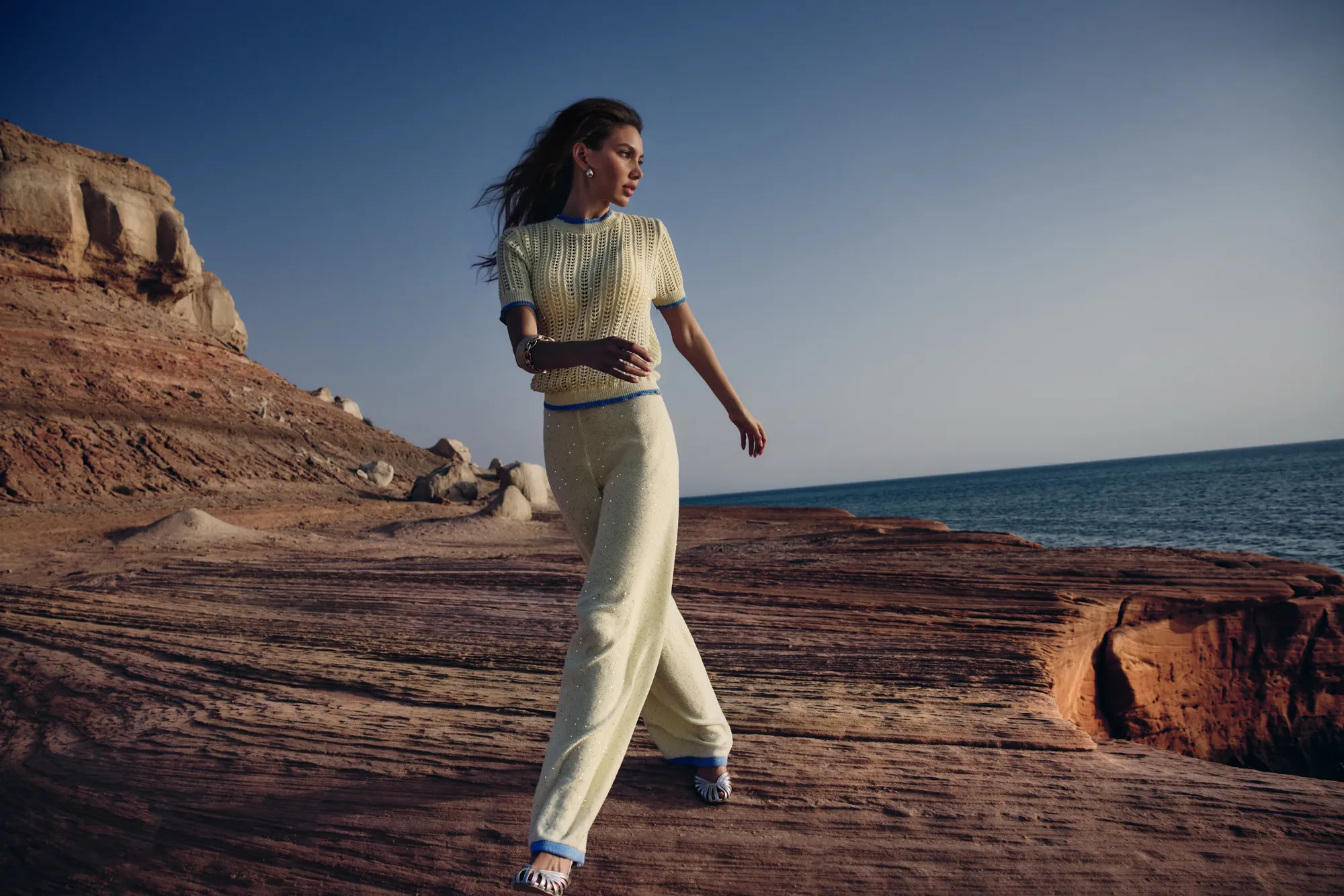 Model walking a red-rock shelf above the sea in a pale-lemon ribbed knit set