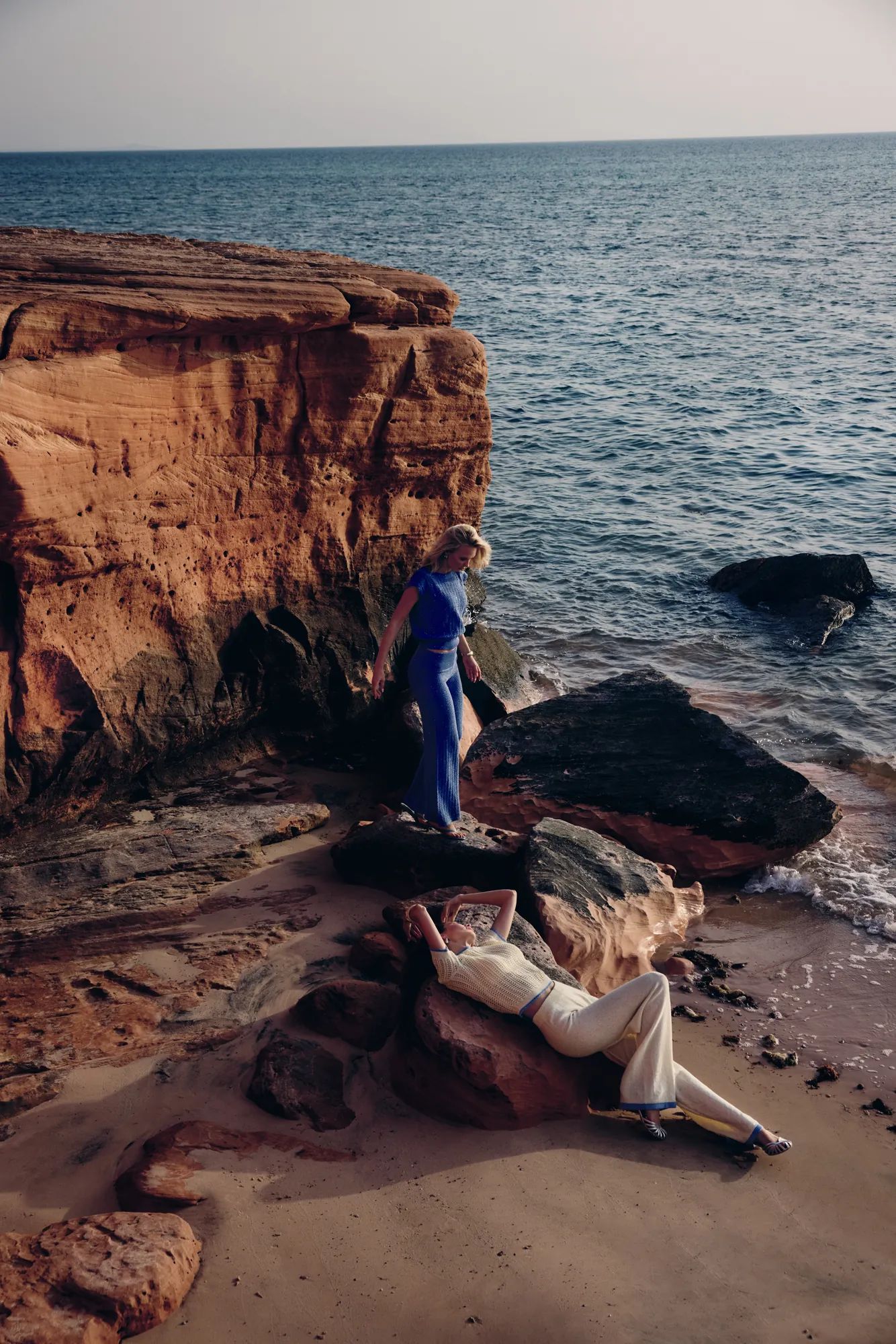 Model in a cobalt-blue knit set standing on daylit coastal rocks above the sea, a second model reclining on the sand below