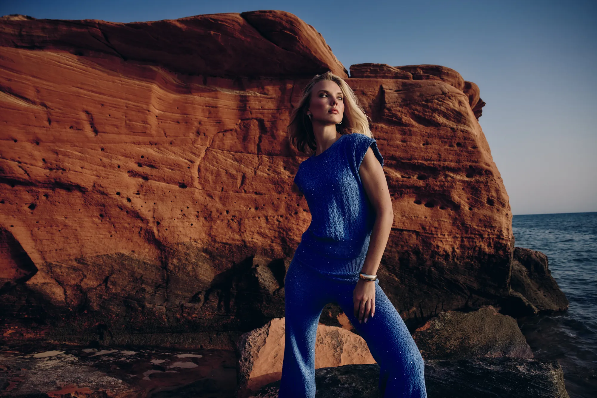 Model in a cobalt sparkle-knit set standing before a monumental red rock as dusk falls over the sea