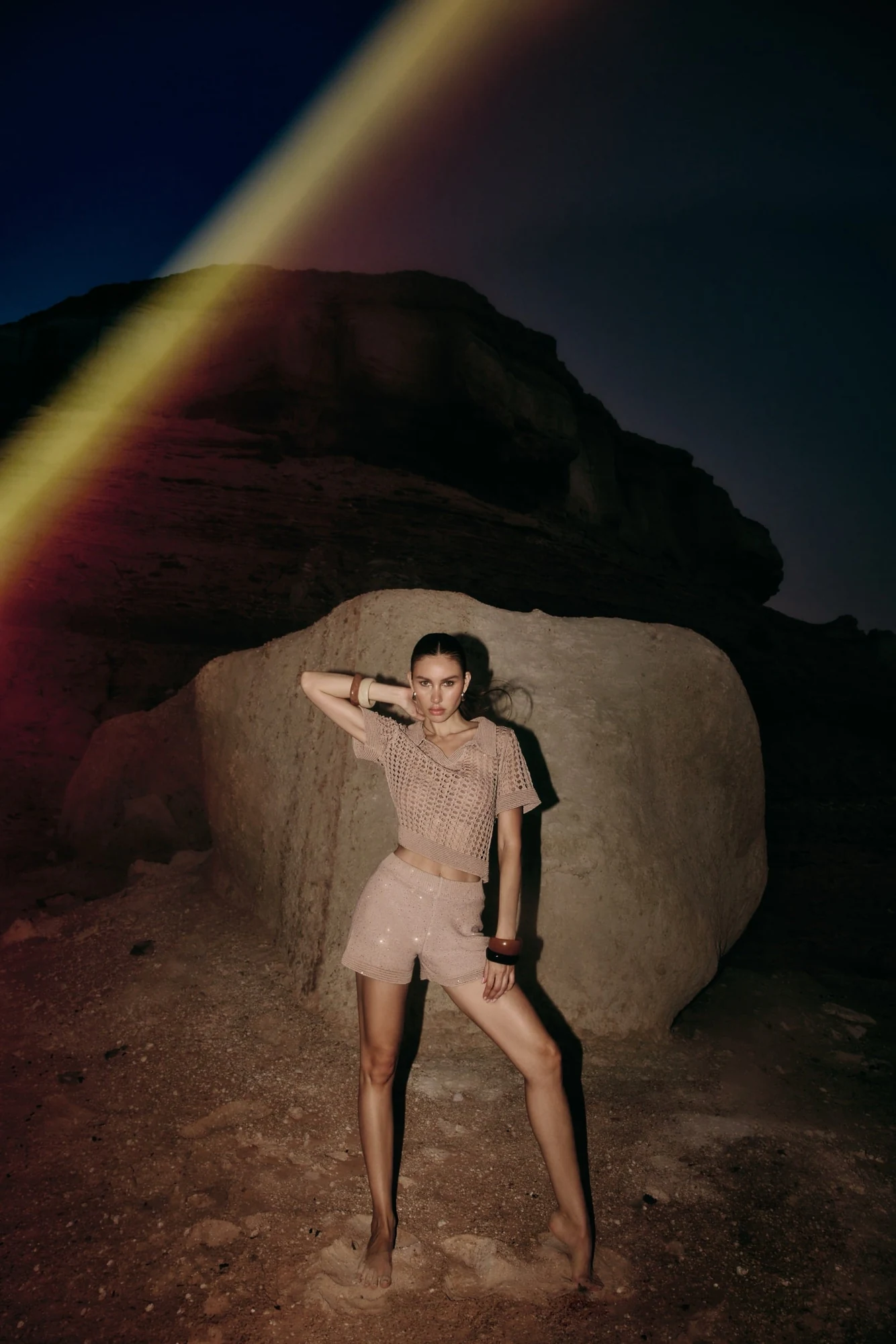 Model in a knit set standing before a vast moonlit rock at night, a rainbow lens flare arcing across the sky