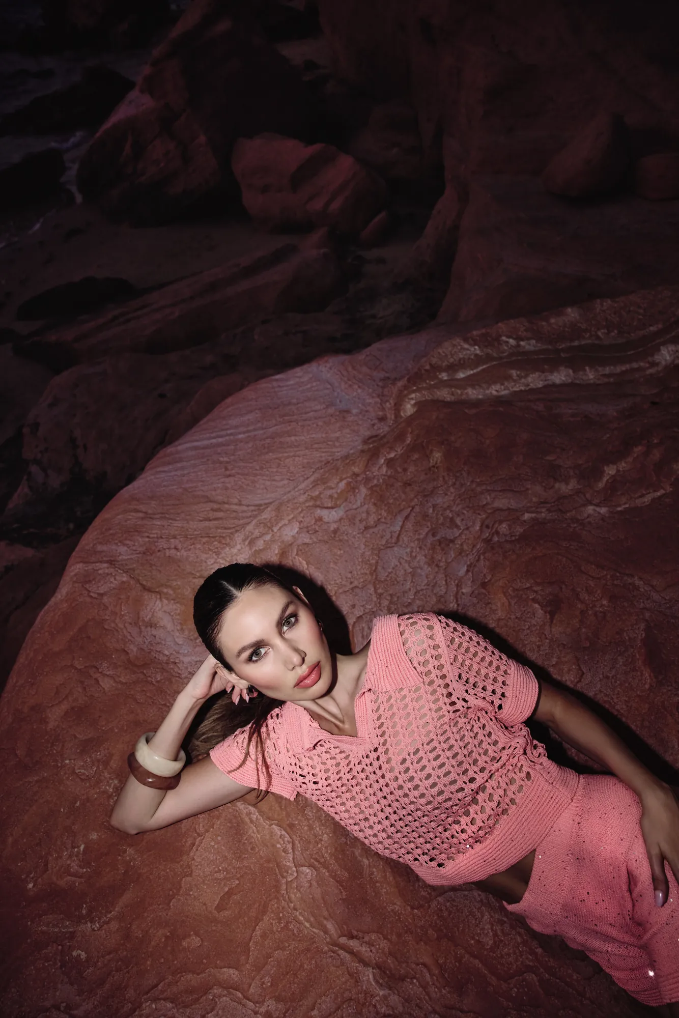 Model in a coral crochet polo and sparkle shorts reclining on a red rock shelf, seen from above