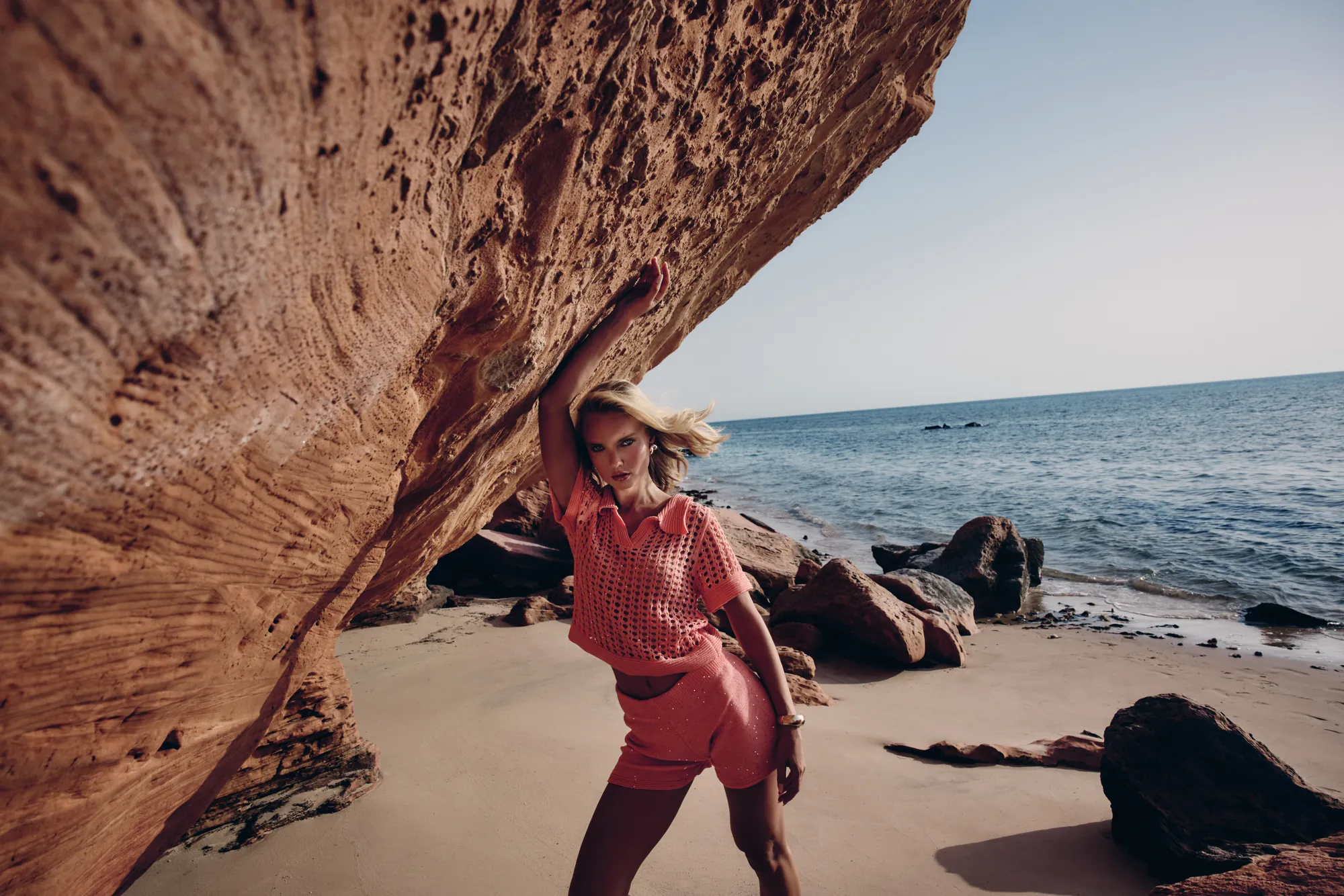 Model in a coral crochet-knit set leaning into a wind-carved rock overhang on a deserted beach