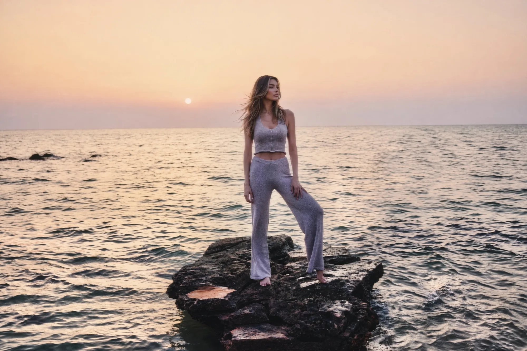 Model in a grey knit set standing barefoot on a sea rock, the sun setting over the water behind her