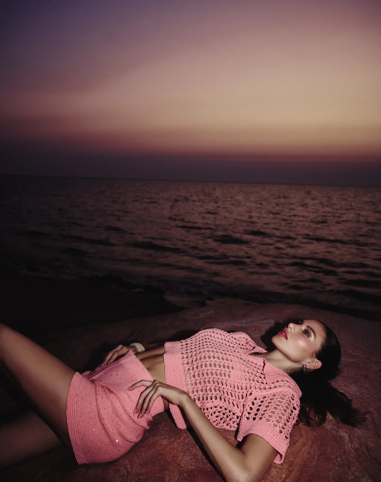 Model in a coral crochet set lying back on red rock above the sea as the sun sets